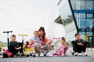 Young stylish mother with four kids outdoor. Sports family spend free time outdoors with scooters and skates. photo