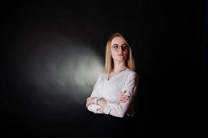 Studio portrait of blonde businesswoman in glasses, white blouse and black skirt against dark background. Successful woman and stylish girl concept. photo