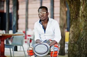 Stylish african american man in white shirt and colored pants with hat and glasses posed outdoor. Black fashionable model boy. photo