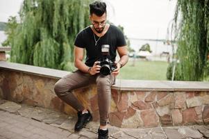 Impresionante y hermoso fotógrafo de hombre macho con barba árabe alta con gafas y camiseta negra con cámara profesional en las manos. foto