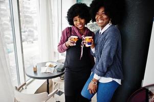 dos mujeres afroamericanas de pelo rizado usan suéteres con tazas de té posadas en el café interior. foto