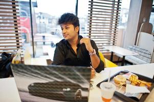 Stylish indian man sitting at fast food cafe against his laptop. photo