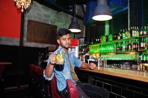 Portrait of handsome successful bearded south asian, young indian freelancer in blue jeans shirt sitting in night club against bar counter with cocktail and having a rest. photo