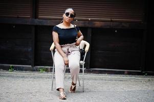 elegante mujer afroamericana con gafas de sol posadas al aire libre en la silla. foto