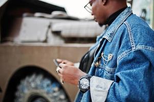 African american man in jeans jacket, beret and eyeglasses, smoking cigar and posed against btr military armored vehicle, with mobile phone at hand. photo