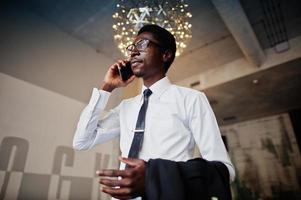 el hombre afroamericano de negocios usa camisa blanca, corbata y anteojos en la oficina hablando por teléfono móvil. foto