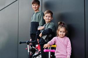 Three kids outdoor against black modern wall. Sports family spend free time outdoors with scooters and skates. photo