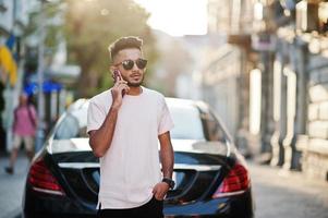 elegante hombre de barba india con gafas de sol y camiseta rosa contra un auto de lujo y hablando por teléfono móvil. rica modelo india posó al aire libre en las calles de la ciudad. foto