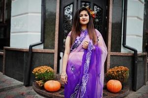 Indian hindu girl at traditional violet saree posed at street against door of restaurant with autumn mood and pumpkins. photo