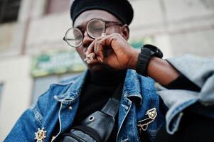 Close up portrait of african american man in jeans jacket, beret and eyeglasses, smoking cigar. photo