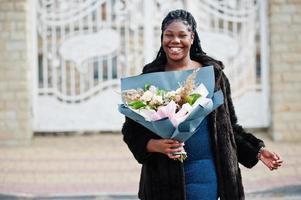 African american dark skinned plus size model posed in a blue shiny dress and black fur coat with bouquet of flowers against large white gates of mansion. photo