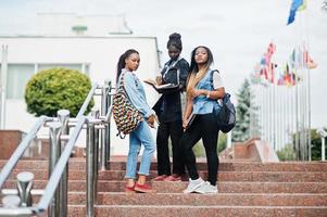 Three african students female posed with backpacks and school items on yard of university and look at book. photo