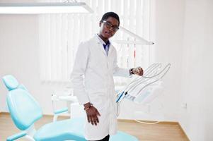 African american male doctor in glasses standing near dentist chair in dental clinic. photo