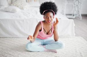 una joven afroamericana con una camiseta rosa escucha música con auriculares en su habitación blanca. foto