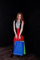 Portrait of an attractive redheaded girl in sleveless jacket, striped blouse and red trousers posing with multicolored packages next to the brick wall. Shopping spree. photo