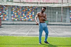 Handsome sexy african american muscular bare torso man at jeans overalls posed at green grass agasinst football gates of stadium field. Fashionable black man portrait. photo