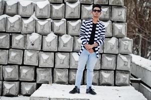 Casual young indian man in stripped sweater and sunglasses posed against stone blocks. photo