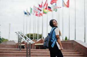 African student female posed with backpack and school items on yard of university, against flags of different countries. photo