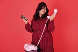 atractiva mujer del sur de Asia vestida de rojo intenso posada en el estudio con fondo rosa y usa un teléfono antiguo con un teléfono móvil moderno. foto