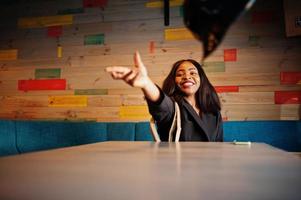 Charming african american woman model in black jacket relaxing in cafe during free time, throwing hat. photo