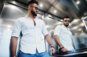 Stylish tall arabian man model in white shirt, jeans and sunglasses posed at elevator inside. photo