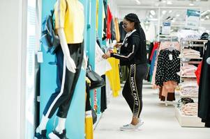 Afican american women in tracksuits and sunglasses shopping at sportswear mall against shelves. She choose yellow t-shirt. Sport store theme. photo