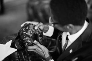 African american photographer paparazzi man wear on black suit and glasses sitting at office with camera and working behind laptop. photo