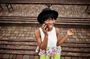 Amazing african american model woman in green pants and black hat posed at bench and using smartphone. photo