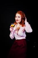 Gorgeous red haired girl in pink blouse and red skirt with glass of wine at hand isolated on black. photo