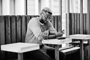 Stylish african american boy on gray sweater and glasses posed at street sitting on table wooden cafe with mobile phone. Fashionable black guy. photo