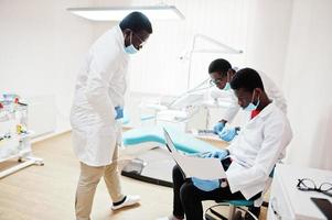 tres médicos afroamericanos trabajando con una laptop, discutiendo con colegas en la clínica dental. foto