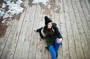 una joven usa una sudadera verde larga, jeans y sombreros negros sentados en un piso de madera con nieve. foto