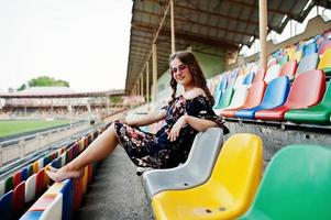 retrato de una joven hermosa vestida y gafas de sol sentada en las tribunas del estadio. foto