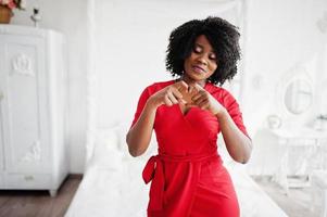 Modelo afroamericano de moda con vestido rojo de belleza, mujer sexy posando vestido de noche en una habitación blanca vintage y muestra el corazón con las manos. foto