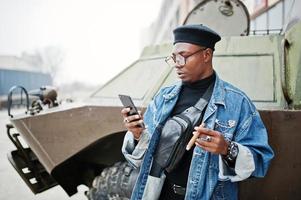 African american man in jeans jacket, beret and eyeglasses, smoking cigar and posed against btr military armored vehicle, with mobile phone at hand. photo