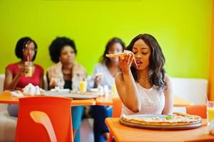 African woman with pizza sitting at restaurant against dark skinned girls. photo