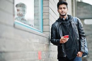 hombre asiático elegante e informal con chaqueta de cuero negro, auriculares con teléfono móvil rojo en las manos posado en la calle. foto