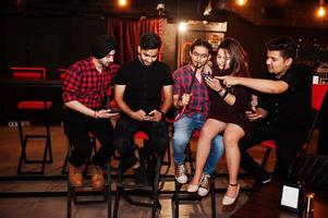 Group of indian friends sitting at lounge bar, having fun and rest, smoking hookah and looking on mobile phones. photo