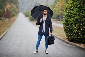 hombre de barba árabe alto de moda vestido con abrigo negro con paraguas y estuche posado en el día de la lluvia. foto