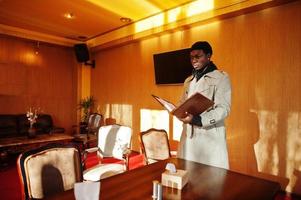 Handsome african american man posing  inside room with sunlight shadows in black hat and beige coat. photo