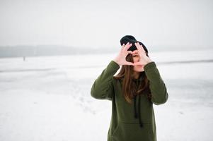 chica divertida vestida con sudadera verde larga, jeans y sombreros negros, en el lago congelado en el día de invierno, mostrando el corazón en los dedos. foto