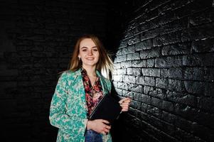 chica rubia con estilo en chaqueta y jeans con diario de cuaderno en las manos contra la pared de ladrillo negro en el estudio. foto