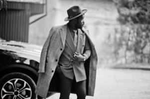 elegante modelo de hombre afroamericano con abrigo gris, corbata de chaqueta y sombrero rojo contra un camión suv negro. foto en blanco y negro.