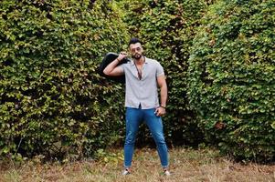 el hombre de barba árabe alto de moda usa camisa, jeans y gafas de sol posados en el parque y sostiene la caja de la bolsa. foto