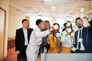 Team of multicultural young people pointing on glass with colorful paper notes. Diverse group of male and female employees in formal wear using stickers. photo