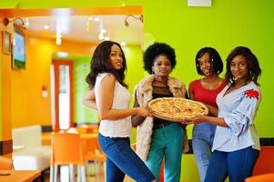 cuatro jóvenes africanas en un restaurante de colores brillantes sostienen una bandeja de madera con pizza en las manos. foto