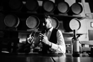 Handsome well-dressed arabian man with glass of whiskey and cigar posed at pub. photo