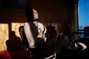 Handsome african american man posing  inside room with sunlight shadows in black hat and beige coat. photo