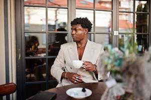 Stylish afro man in beige old school suit sitting on cafe with cup of coffee. Fashionable young African male in casual jacket on bare torso. photo