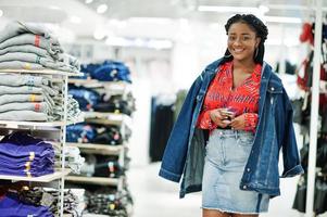 Modish african american woman in  red shirt and jeans skirt with jacket posed at clothes store. It's time for shopping. photo